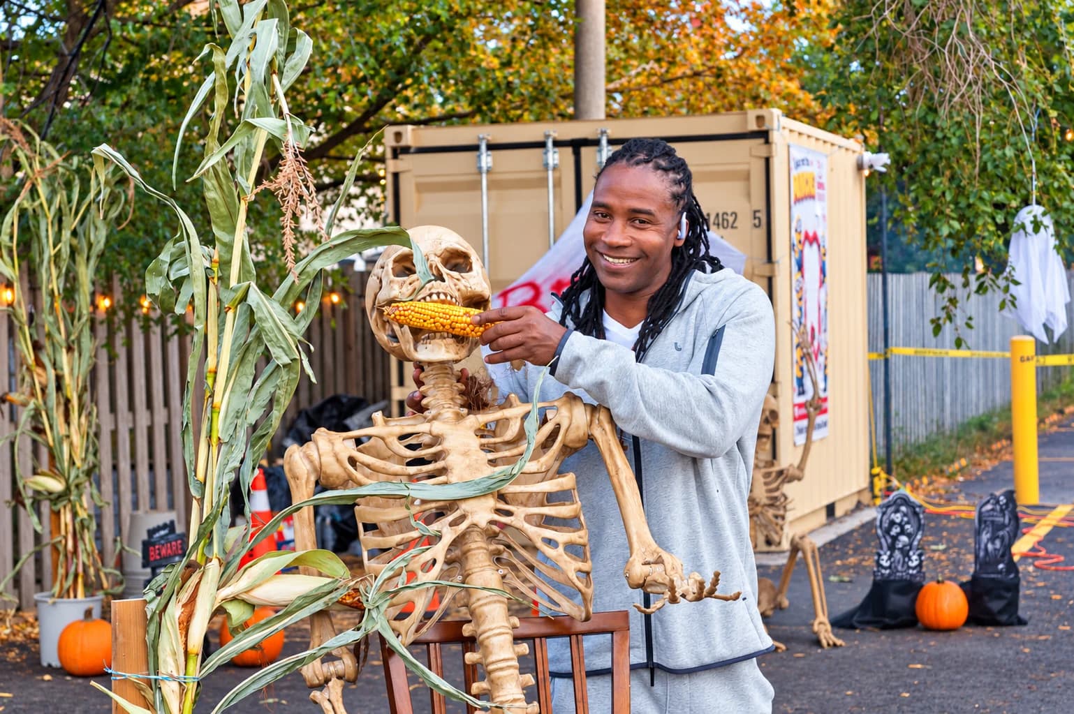 A smiling man with braided hair holds a corn cob beside a seated skeleton decoration.