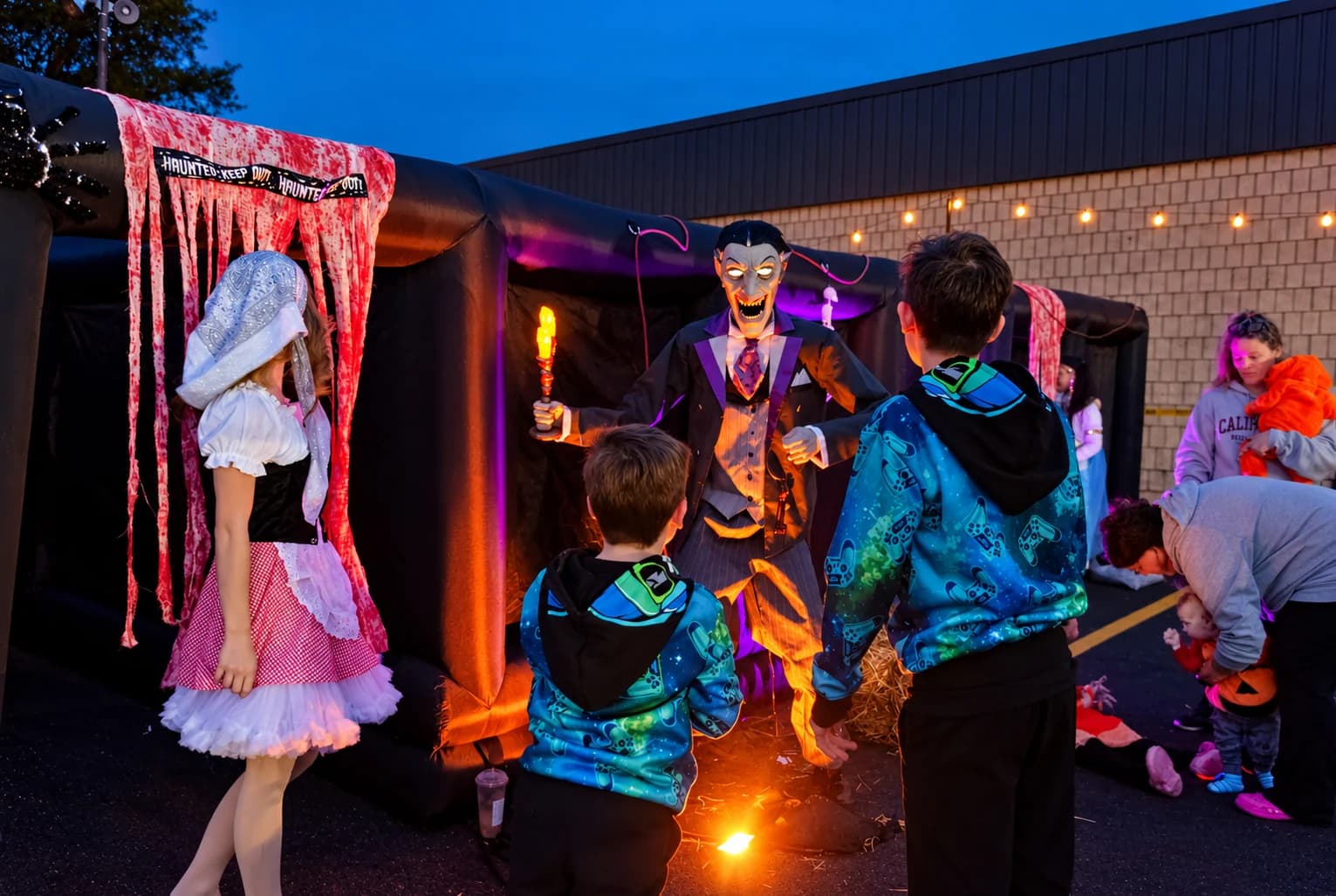 Children and a costumed host stand beside an illuminated vampire prop and the haunted-house entrance.